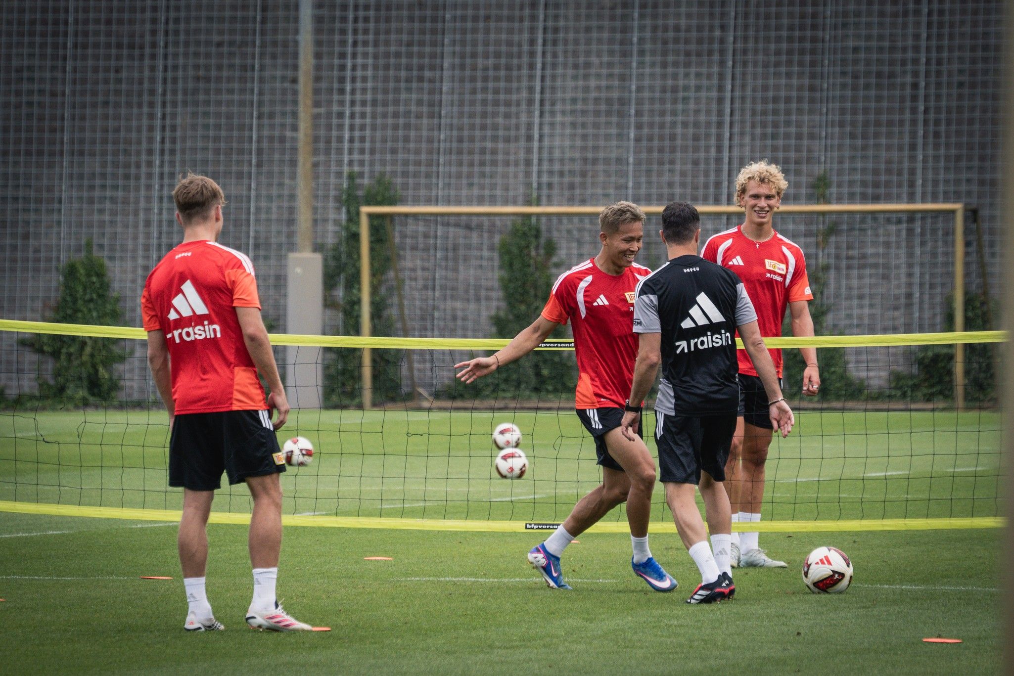 Vier Fußballspieler in roten und schwarzen Trikots trainieren auf einem grünen Platz mit Toren im Hintergrund.