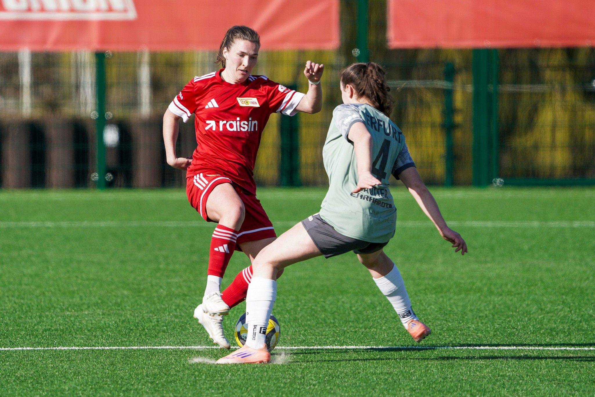 Zwei Fußballspielerinnen in roten und grauen Trikots kämpfen um den Ball auf einem Kunstrasenplatz.