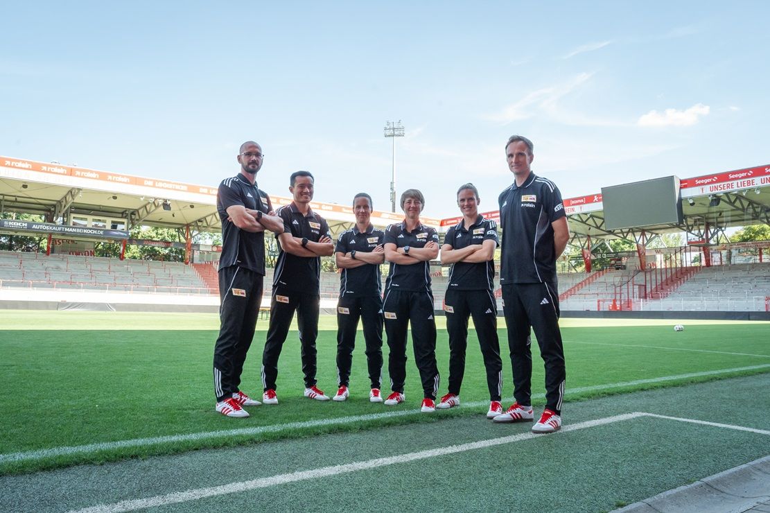 Sechs Personen in sportlicher Kleidung stehen auf einem Fußballfeld in einem Stadion, umgeben von Tribünen.