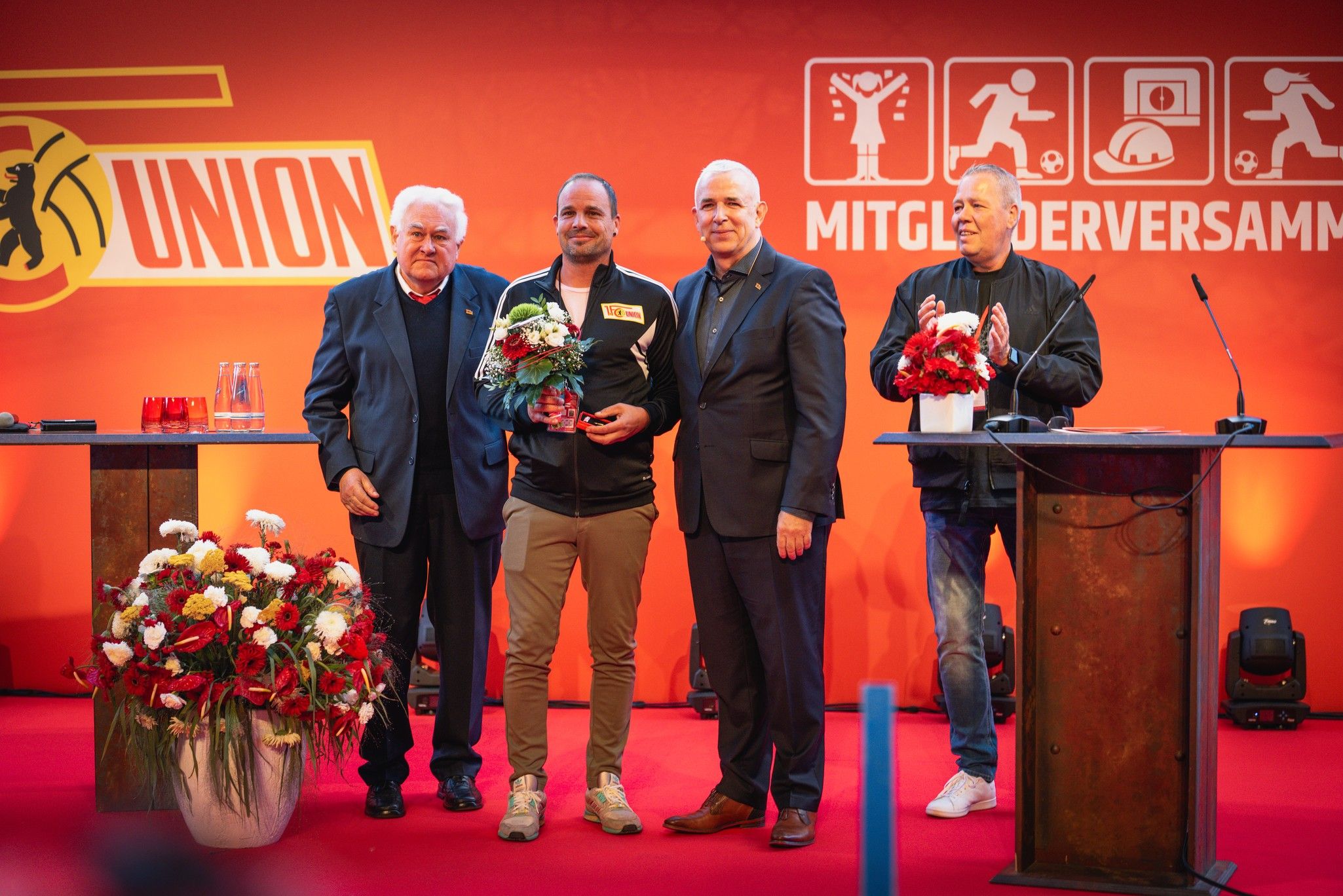 Vier Personen stehen auf einer Bühne, umgeben von roten und weißen Blumenarrangements, bei einer feierlichen Veranstaltung.
