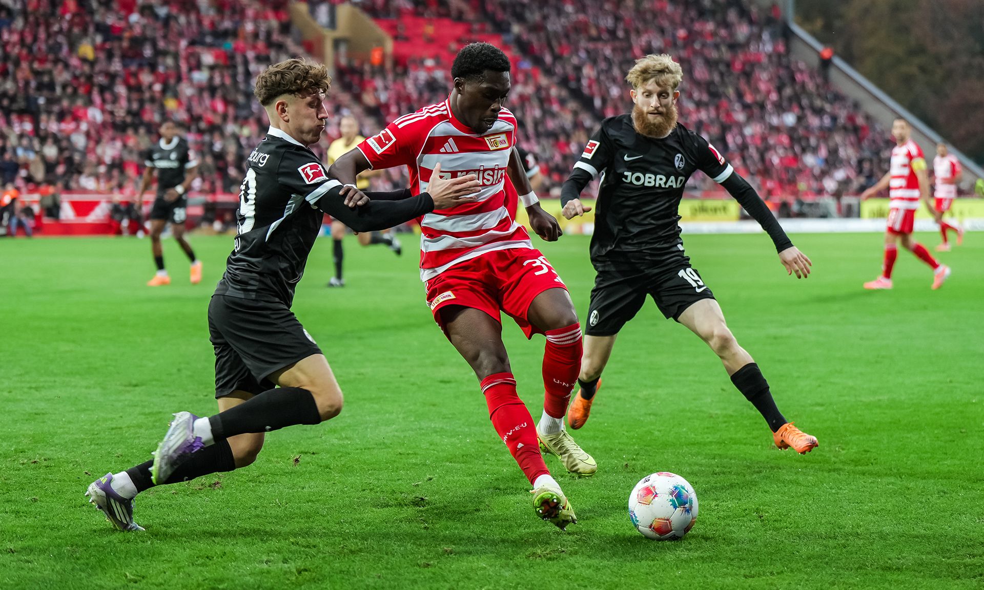 Spielszene aus einem Fußballmatch: Ein Spieler in roter und weißer Kleidung wird von zwei Gegnern in schwarzer Kleidung verfolgt.
