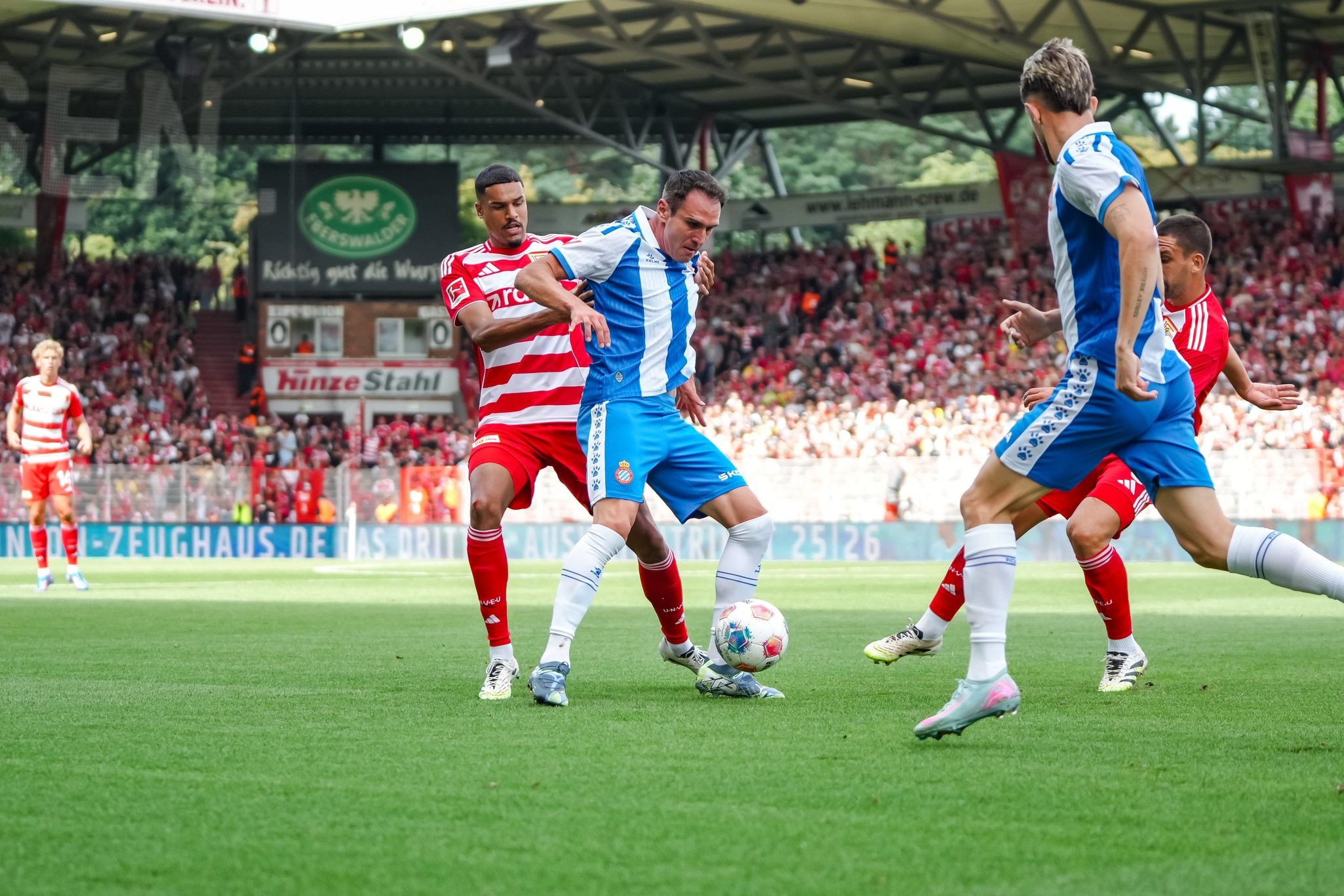 Ein Fußballspiel, bei dem ein Spieler in blauer und weißer Kleidung versucht, den Ball zu dribbeln, während zwei Gegenspieler ihn anpressen.