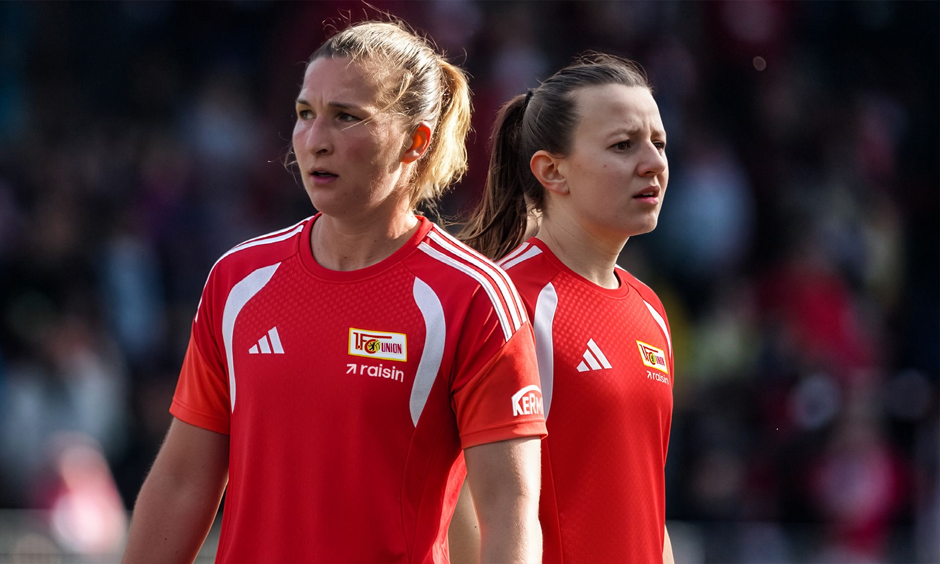 Zwei Fußballspielerinnen in roten Trikots stehen auf dem Spielfeld und blicken ernst in die Ferne.