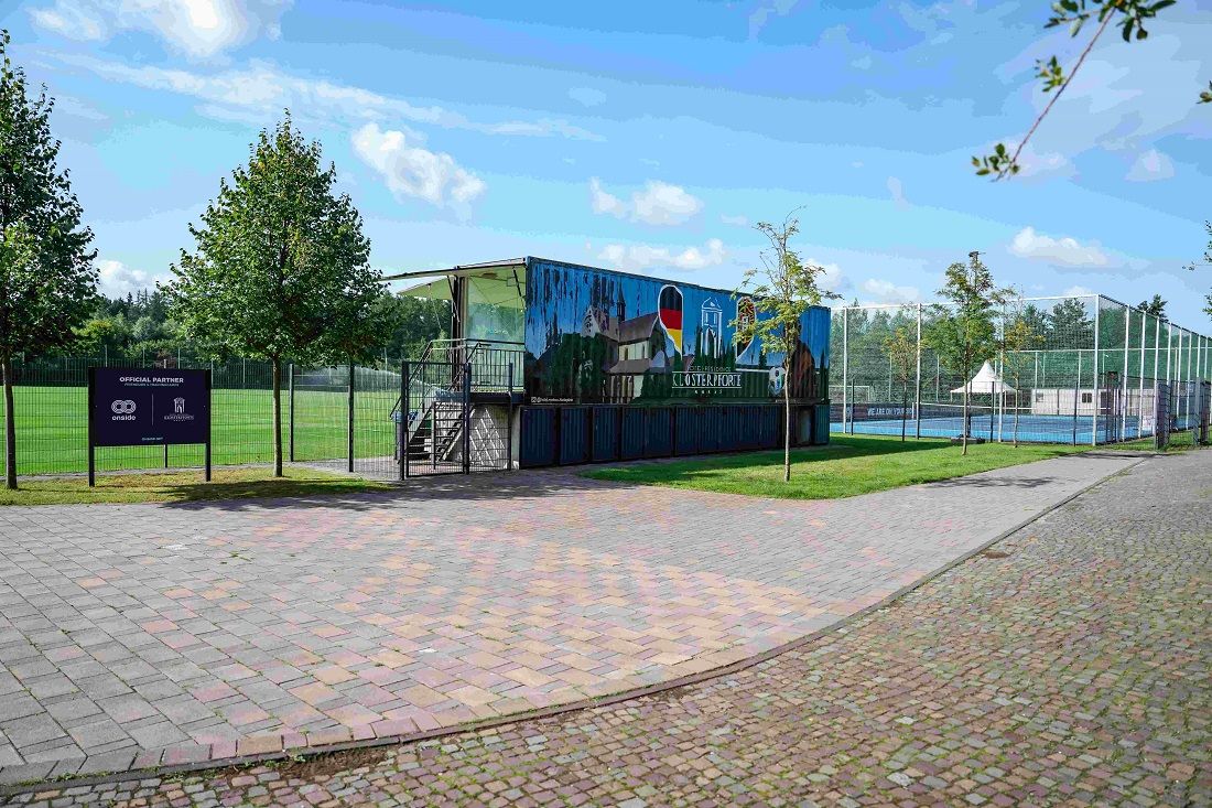 Sportanlage mit Tennisplätzen, einem Sportheim und Bäumen, umgeben von einem gepflasterten Weg und grünen Wiesen.
