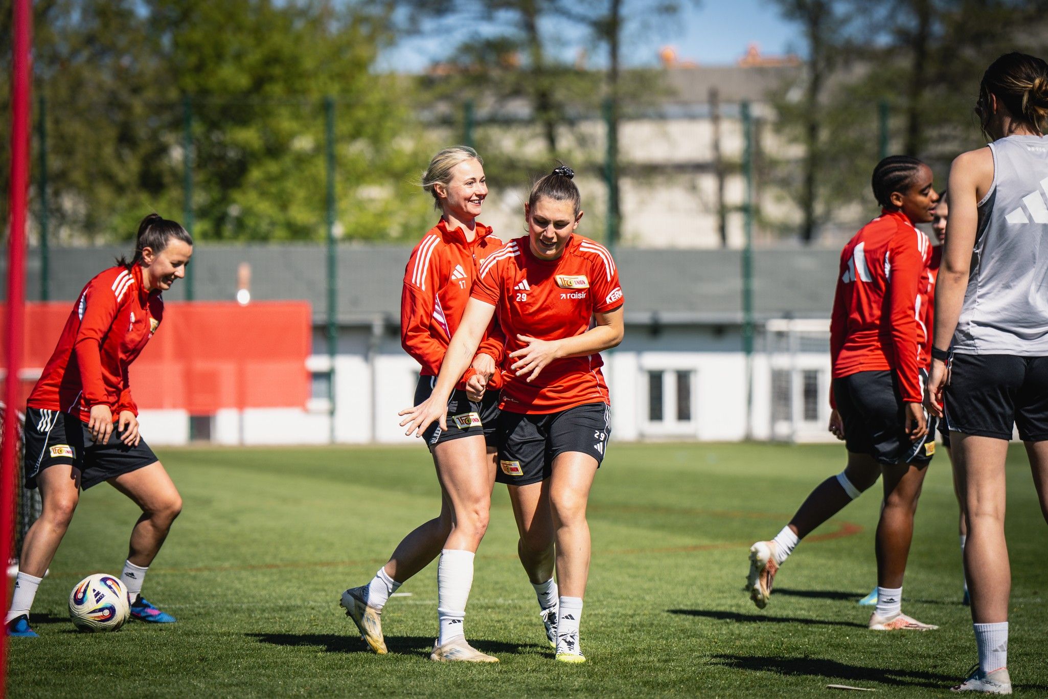 Fünf Frauenfußballerinnen in roten Trikots beim Training auf einem grünen Platz, lachen und interagieren miteinander.