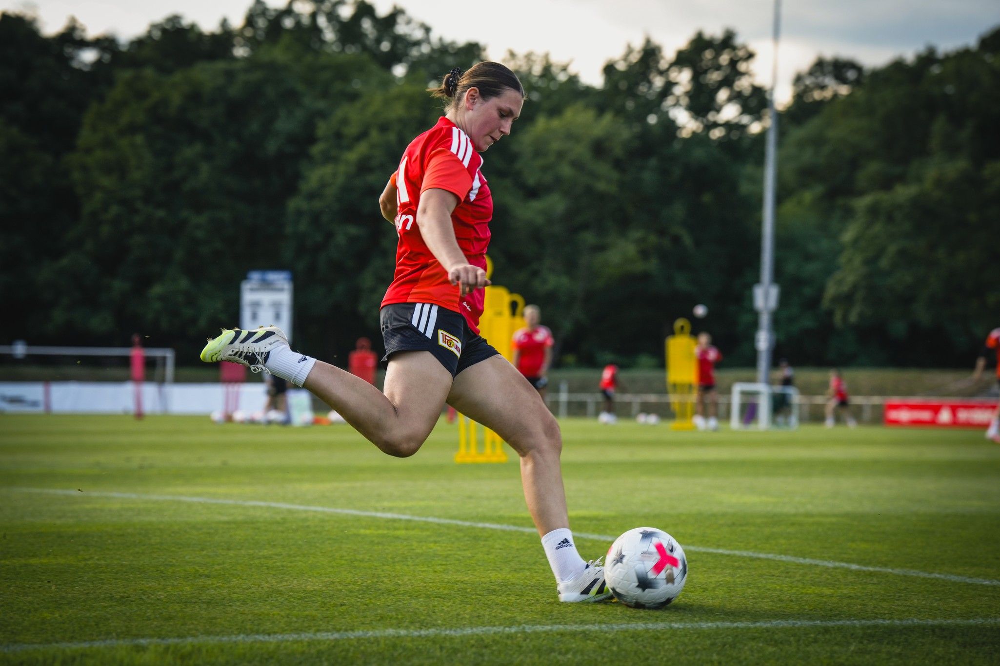 Eine Fußballspielerin in einem roten Trikot tritt den Ball auf einem Trainingsplatz. Im Hintergrund sind weitere Spielerinnen zu sehen.