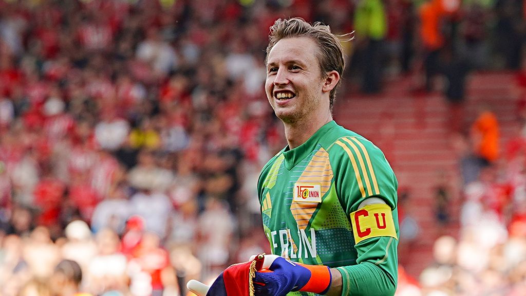 Smiling goalkeeper in green captain's gear, holding a soccer ball in hand, standing in front of cheering crowd.