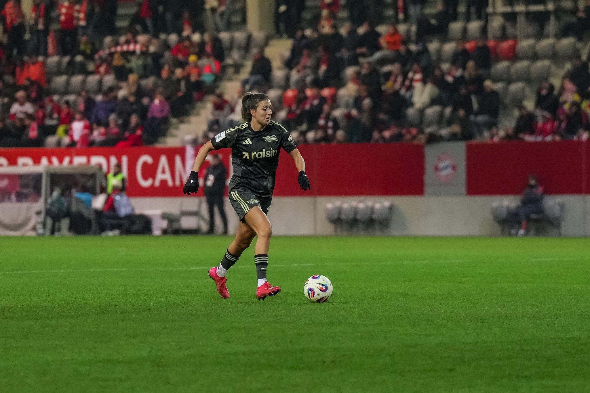 Eine Fußballspielerin in schwarzer Kleidung dribbelt den Ball auf einem grünen Feld während eines Spiels im Stadion.