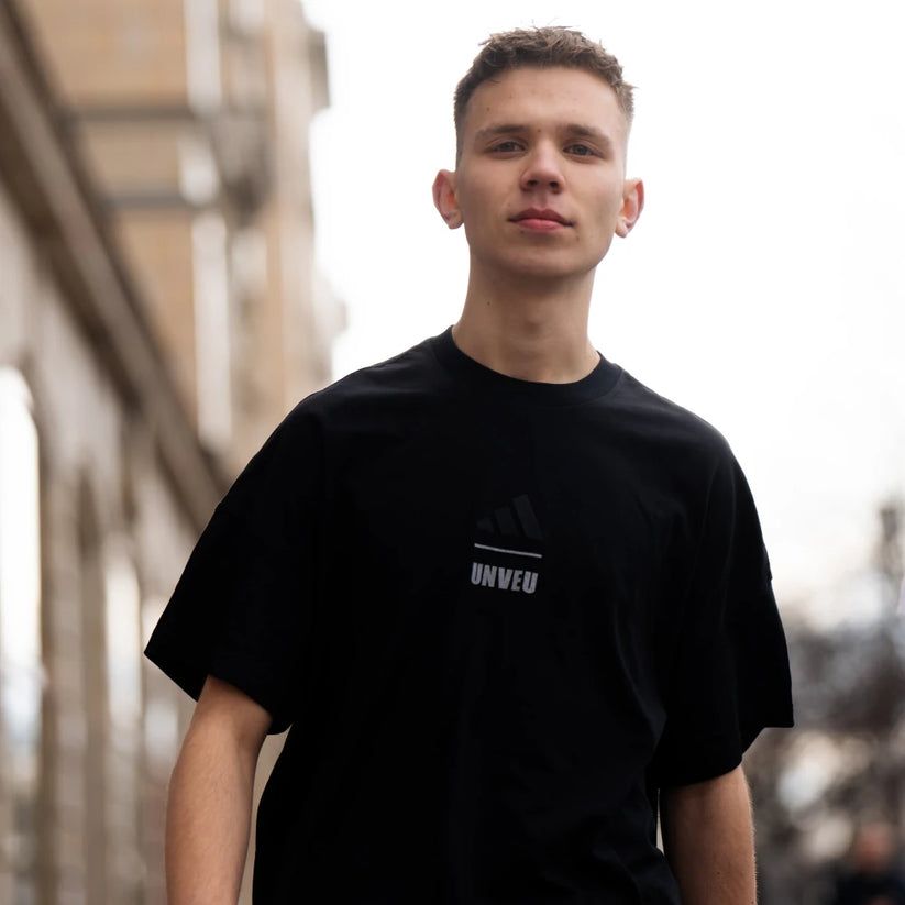 Junger Mann in einem schwarzen T-Shirt mit Adidas-Logo, der in einer städtischen Umgebung posiert.