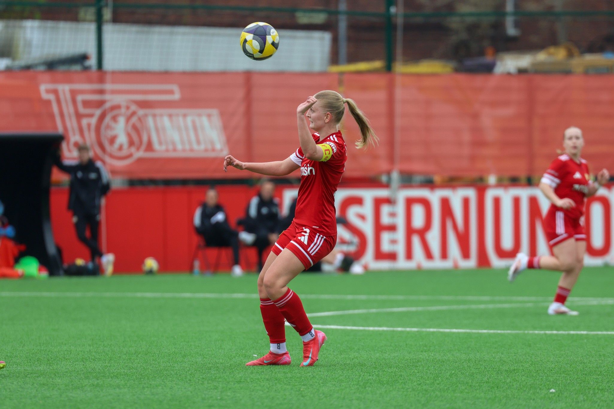Spielerin in rotem Trikot mit einer Ballanpassung auf einem Fußballfeld, im Hintergrund weitere Spielerinnen und Zuschauer.