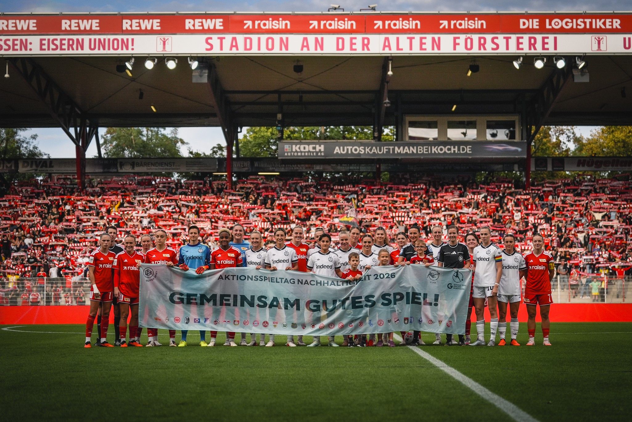 Zwei Fußballmannschaften stehen zusammen auf dem Platz und halten ein Banner mit der Aufschrift „GEMEINSAM GUTES SPIEL!“ hoch.