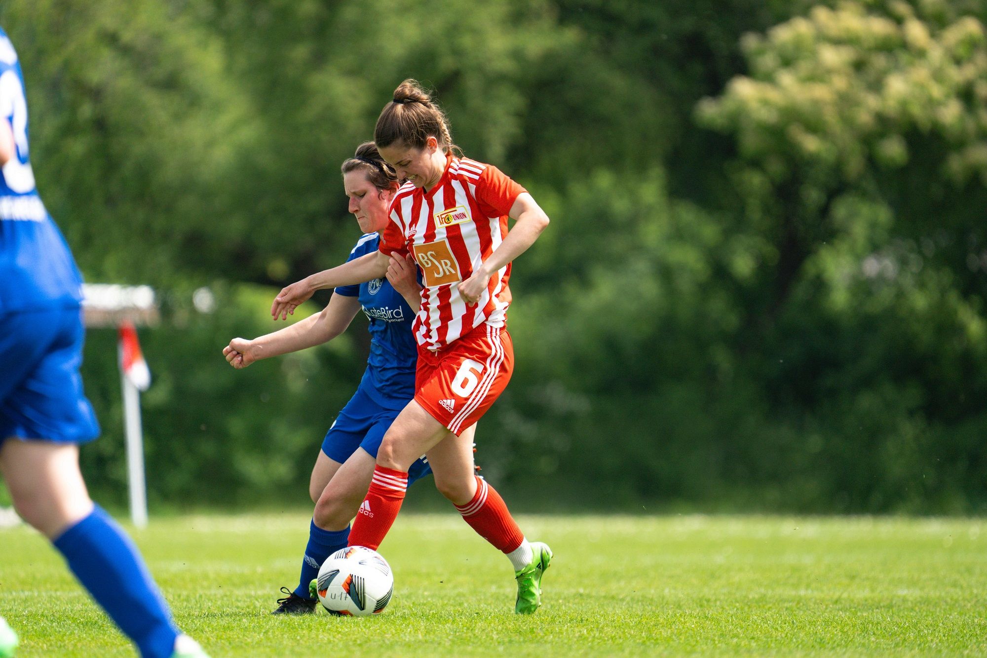 Eine Spielerin in rot-weißem Trikot dribbelt den Ball, während eine Gegnerin in blauem Trikot versucht, ihn zu stoppen.