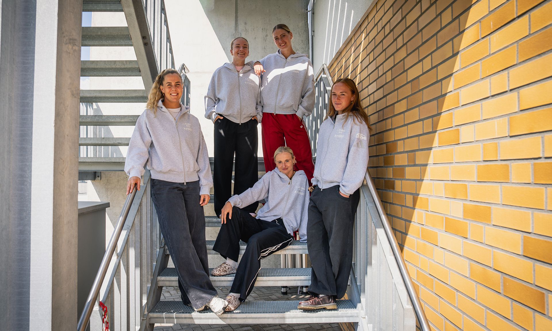 Fünf Personen in gemütlichen grauen Sweatshirts posieren auf einer Treppe vor einer Backsteinmauer.