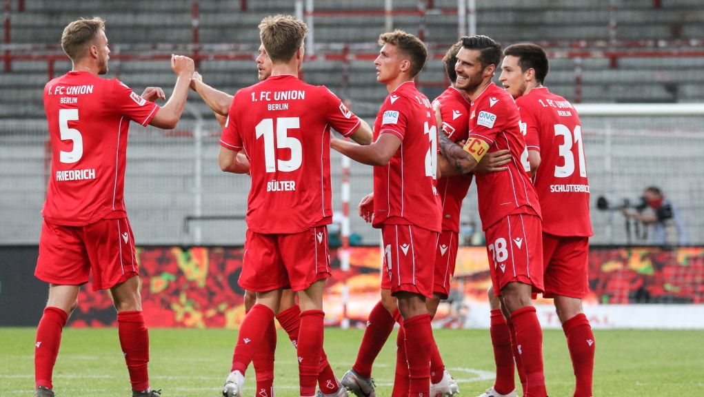 Six soccer players from 1. FC Union Berlin celebrate a goal on the field in red jerseys.