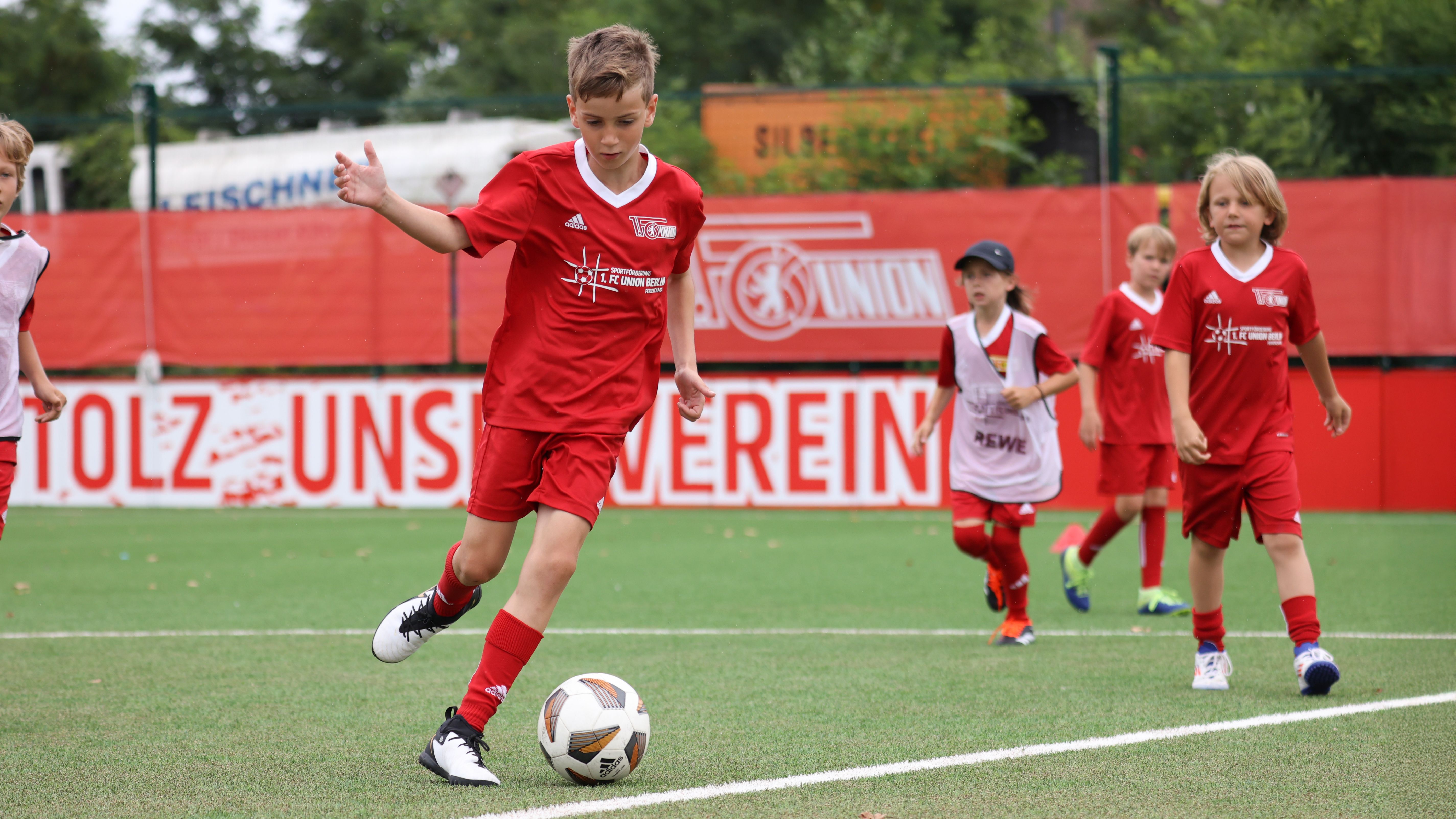 Ein Junge in rotem Fußballtrikot dribbelt einen Ball, während andere Kinder im Hintergrund trainieren.