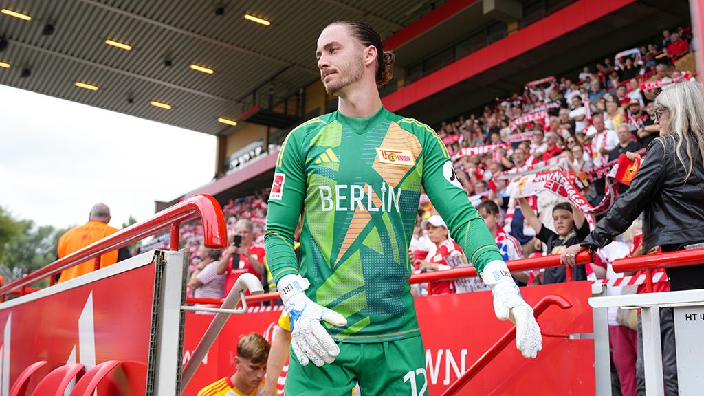 Ein Fußballtorwart in grüner Ausrüstung steht an der Seitenlinie eines Stadions, umgeben von jubelnden Fans.