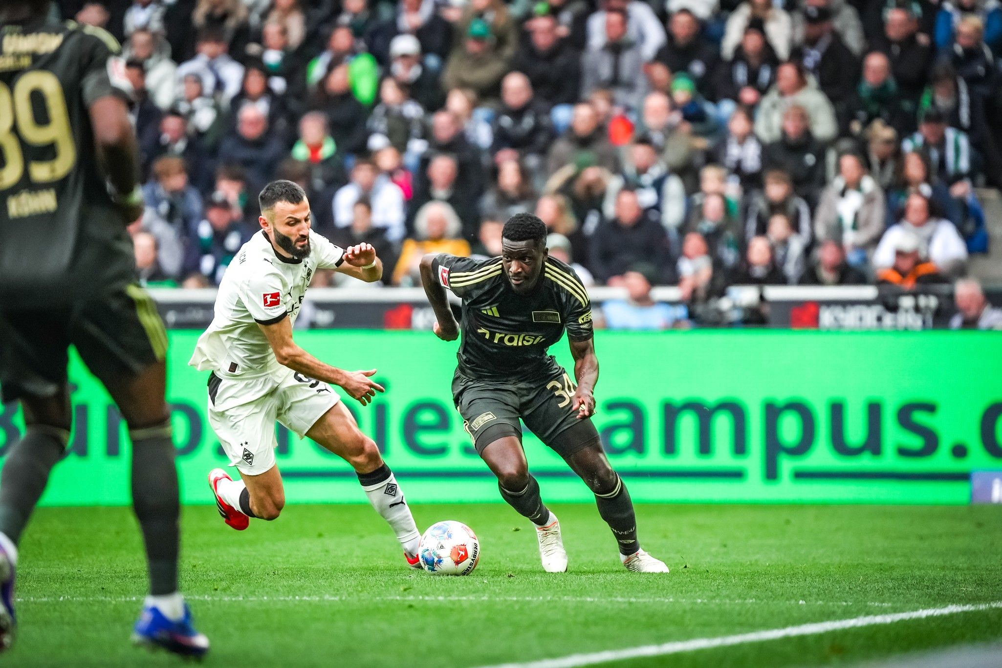 Ein Fußballspieler in dunkler Uniform dribbelt den Ball, während ein gegnerischer Spieler ihn verfolgt. Im Hintergrund Zuschauer.