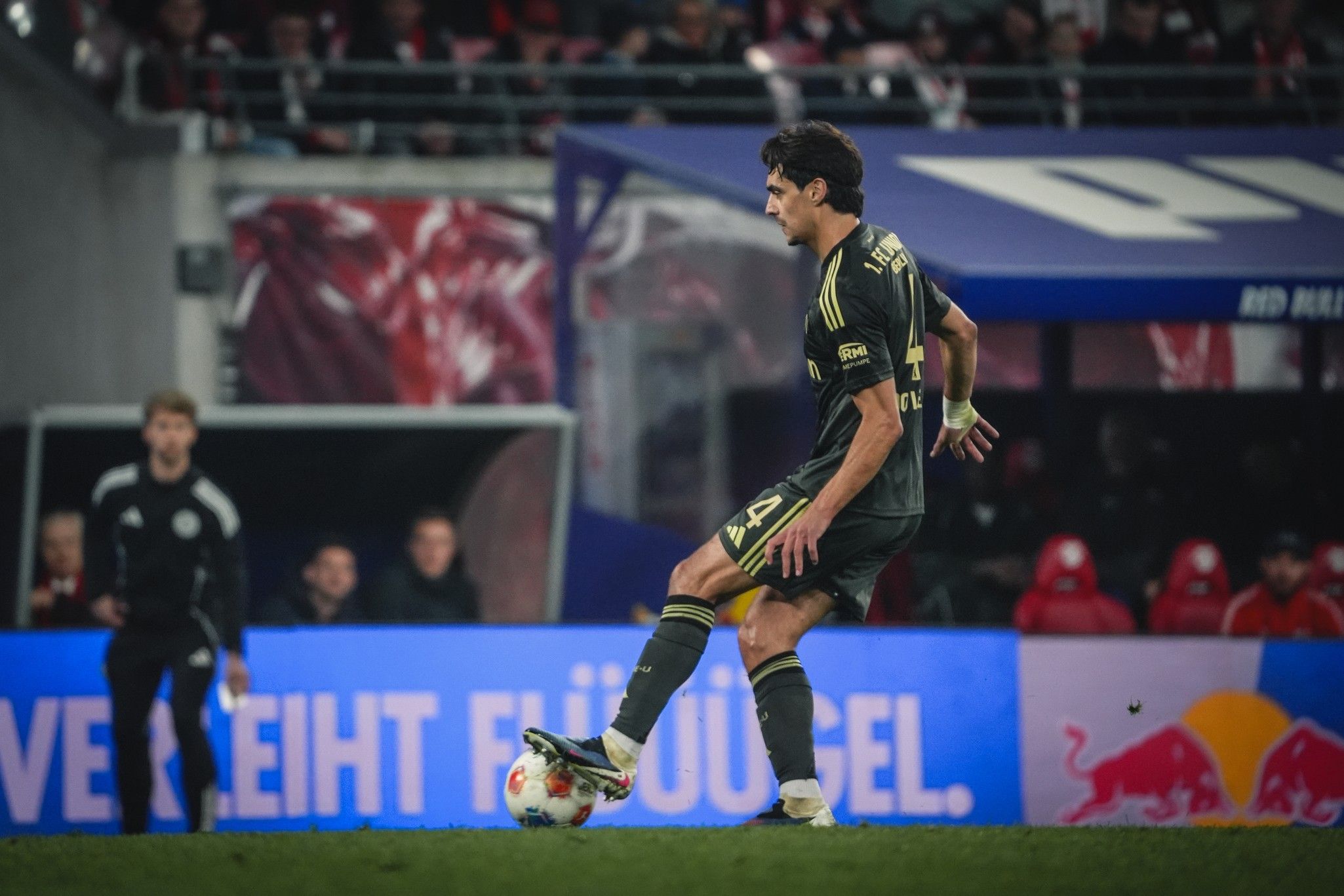Ein Spieler in schwarzem Trikot dribbelt mit dem Ball auf dem Spielfeld, im Hintergrund sind Zuschauer und eine Werbebande sichtbar.