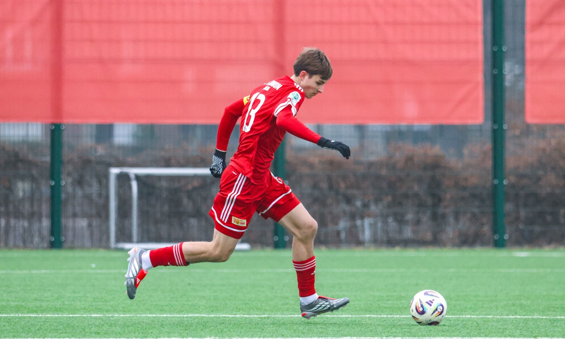 Ein junger Fußballspieler in roter Uniform läuft über das Spielfeld und dribbelt den Ball.