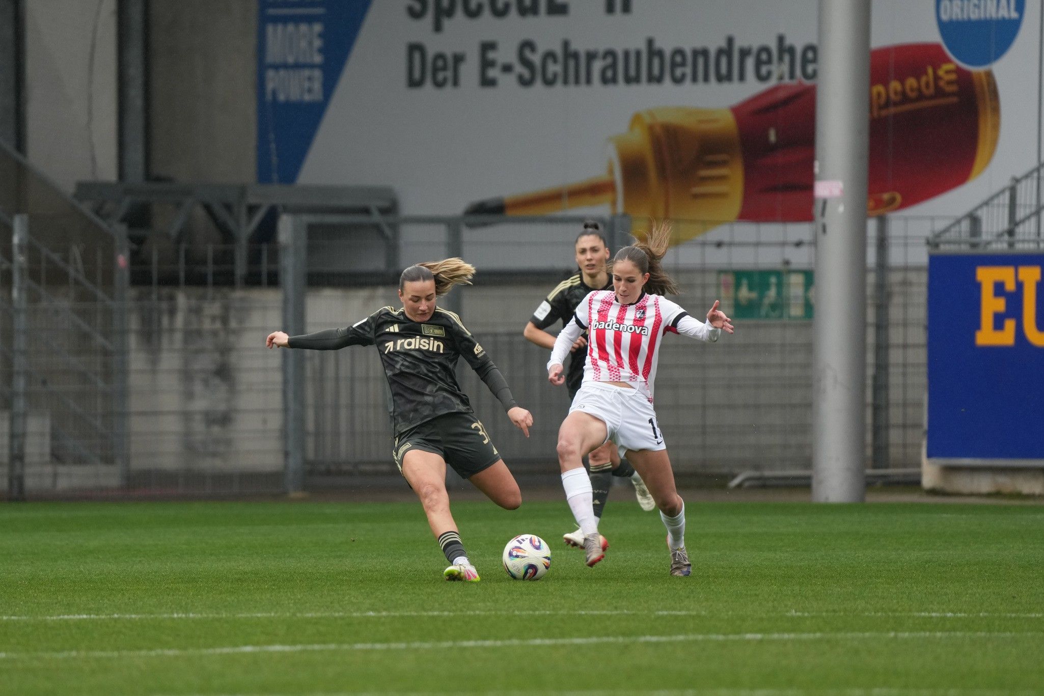 Zwei Fußballspielerinnen kämpfen um den Ball auf einem Spielfeld, während eine Schiedsrichterin im Hintergrund zuschaut.