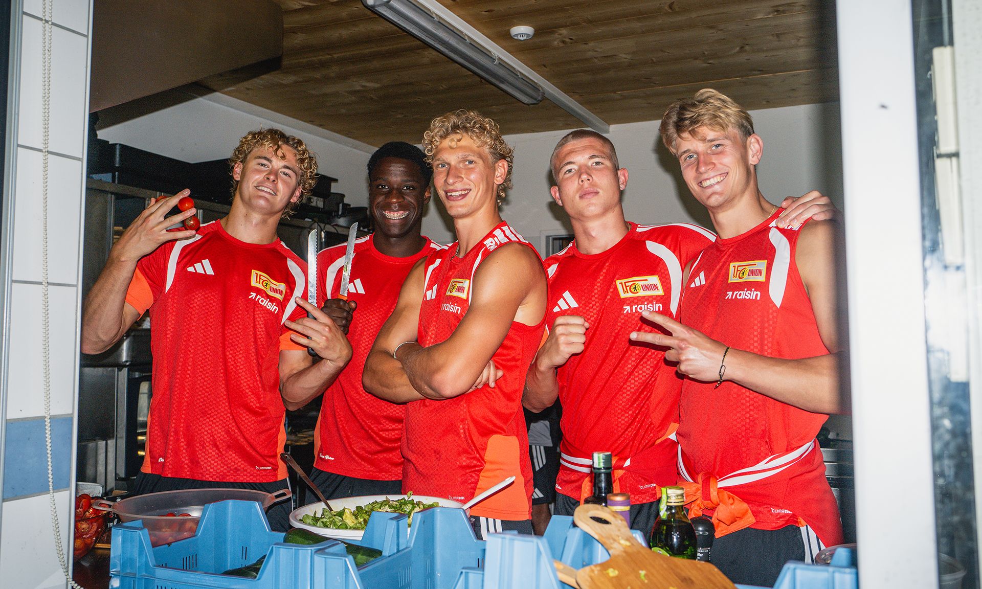 Fünf junge Sportler in roter Sportkleidung posieren lächelnd in einer Küche.