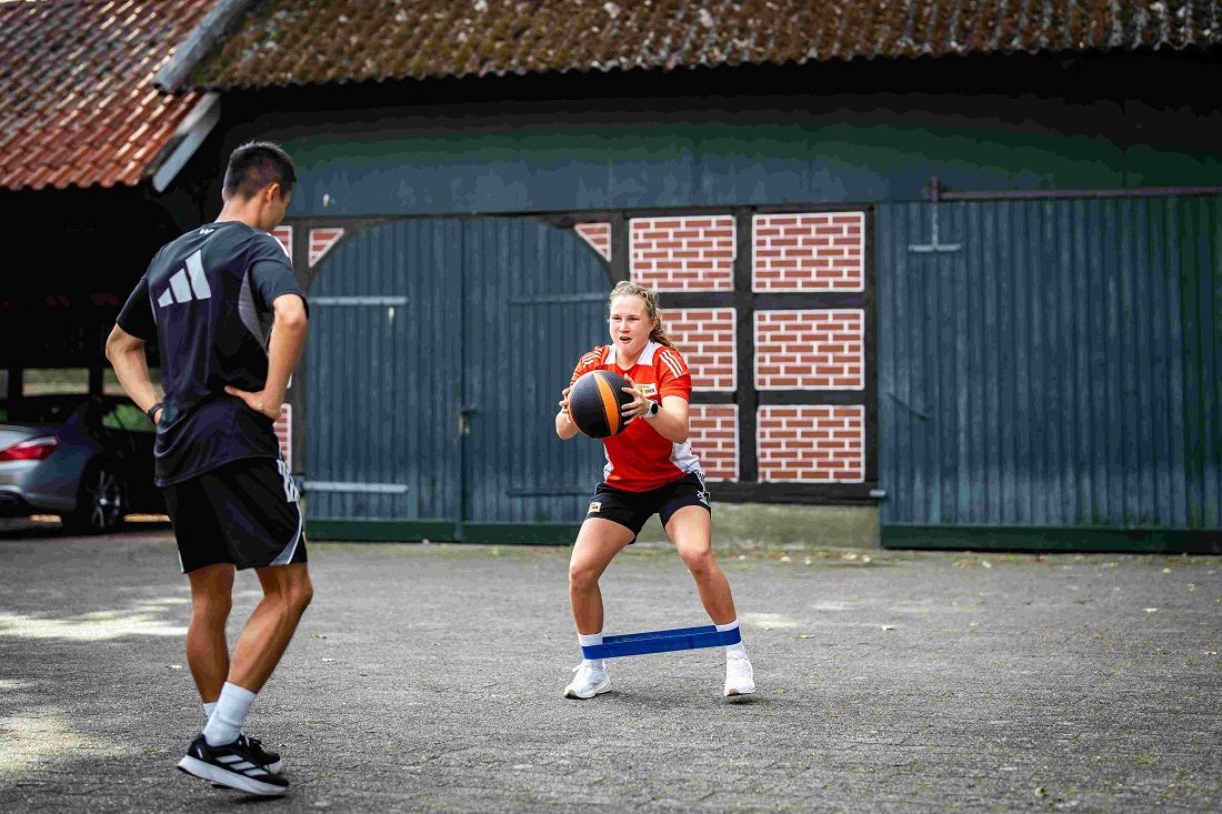 Eine Frau in Sportkleidung übt mit einem Medizinball, während ein Mann sie beobachtet. Beide tragen Sportbekleidung.
