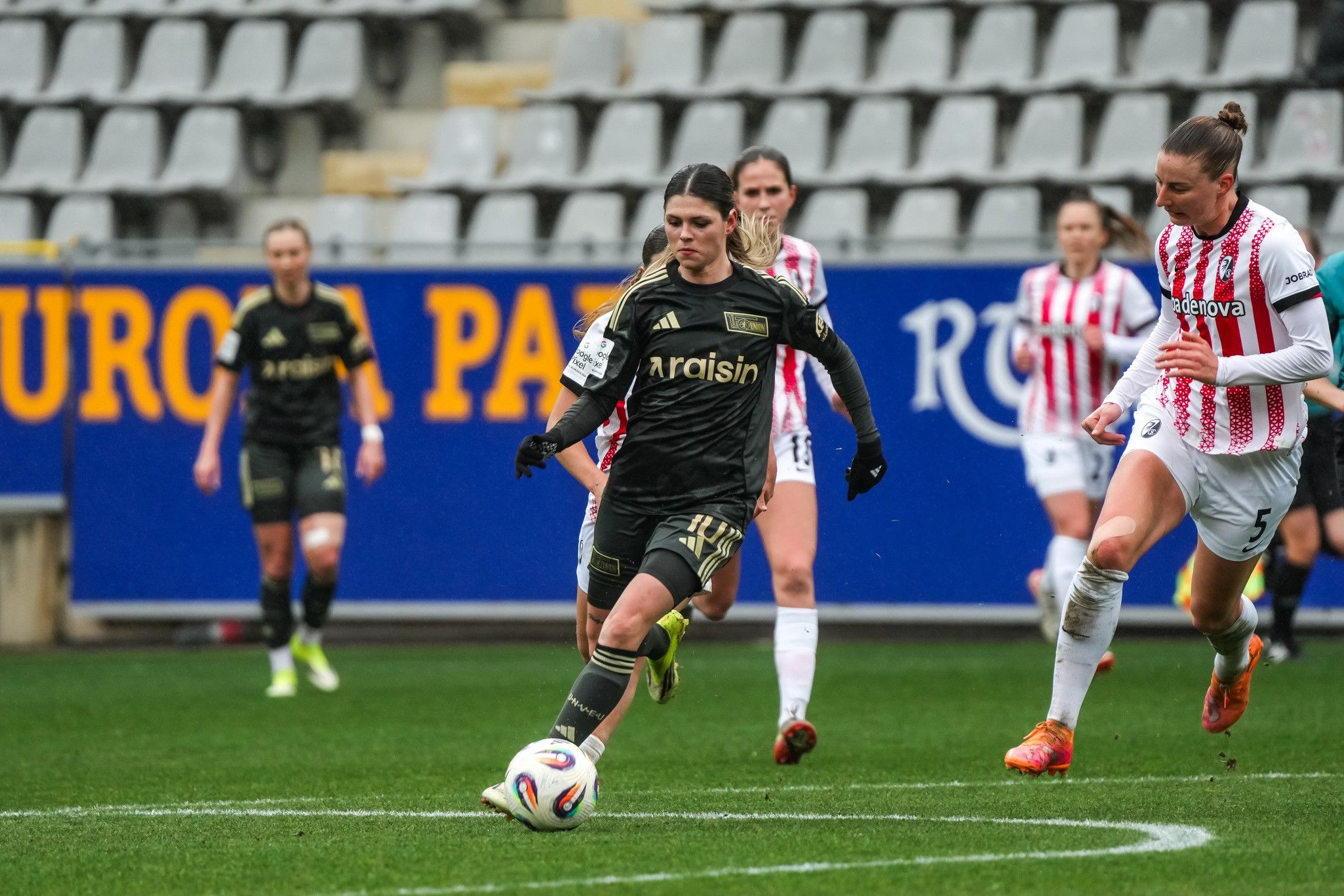 Eine Spielerin in schwarzer Fußballausrüstung läuft mit dem Ball, während sie von einer Gegnerin in weißer und roter Kleidung verfolgt wird.