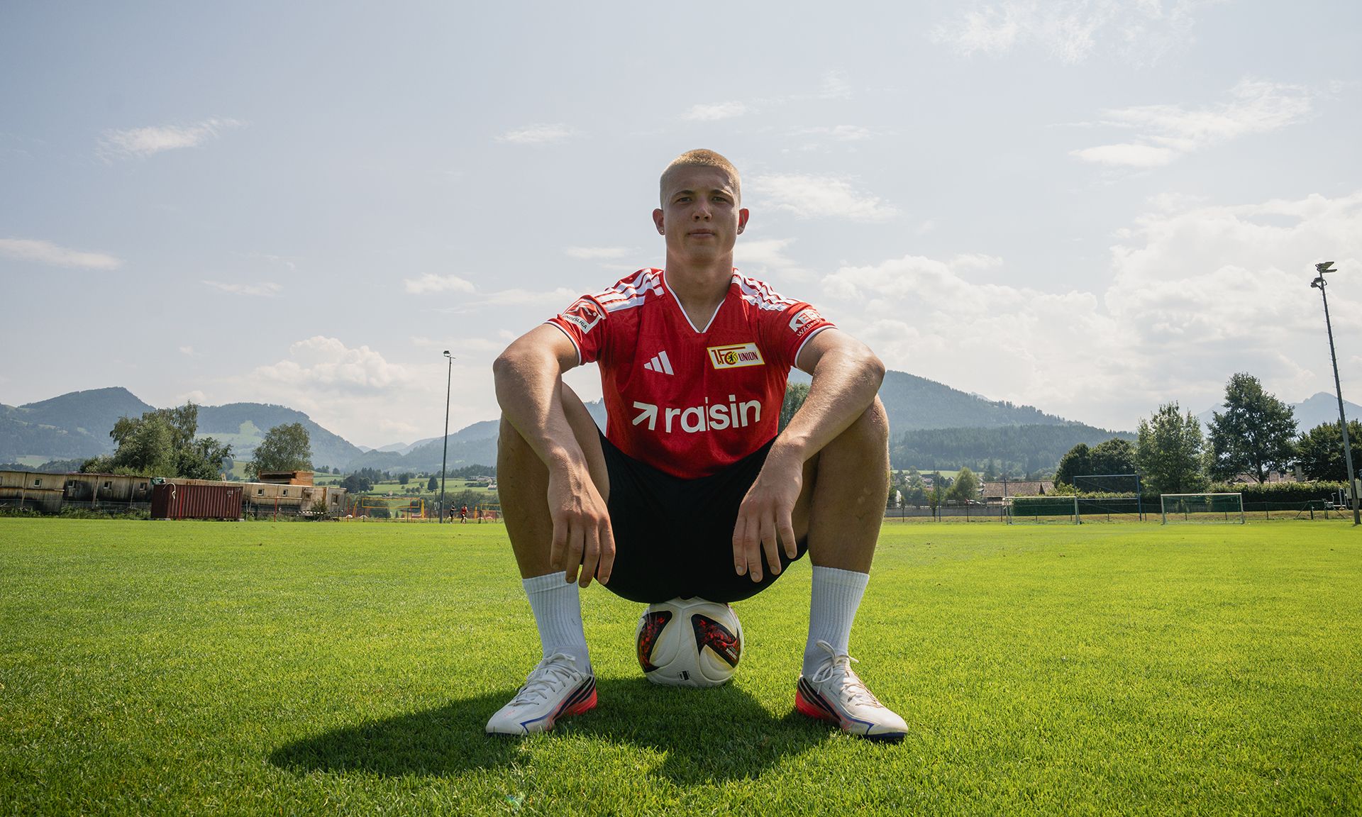 Ein junger Mann sitzt auf einem Fußball, trägt ein rotes Trikot mit dem Schriftzug "rasin" und posiert auf einem grünen Fußballfeld.