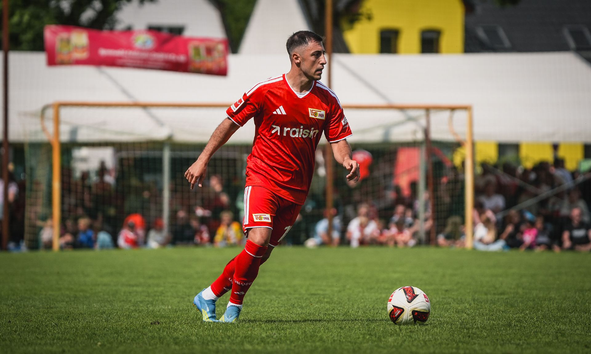 Ein Fußballspieler in rotem Trikot dribbelt den Ball auf einem grünen Spielfeld, umgeben von Zuschauern.