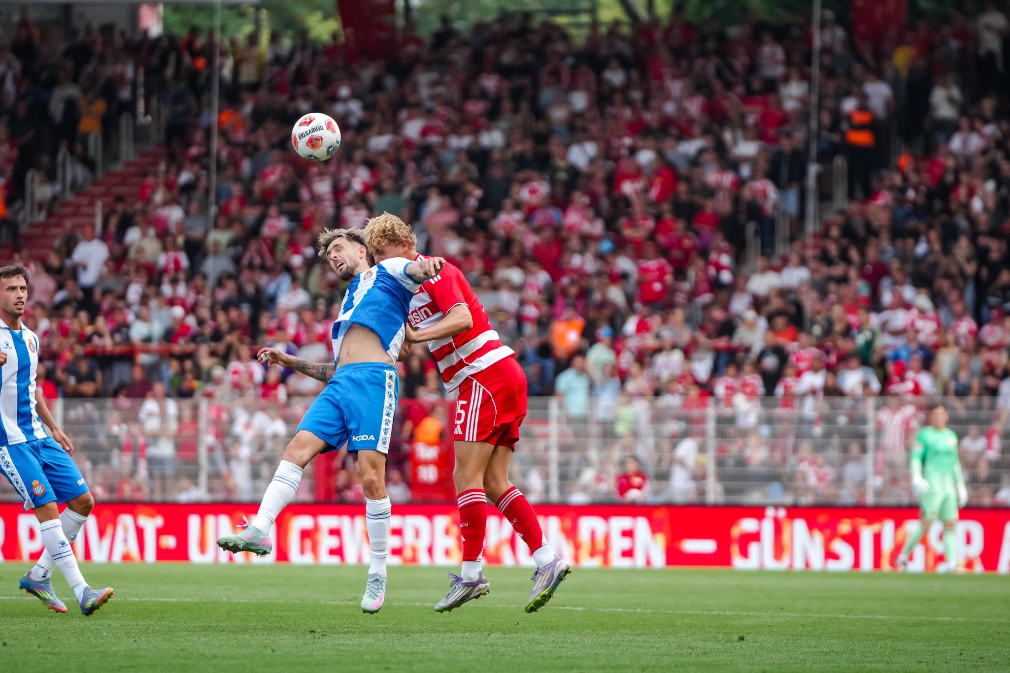 Zwei Fußballspieler im Sprung um einen Ball zu kämpfen, im Hintergrund eine jubelnde Menschenmenge.
