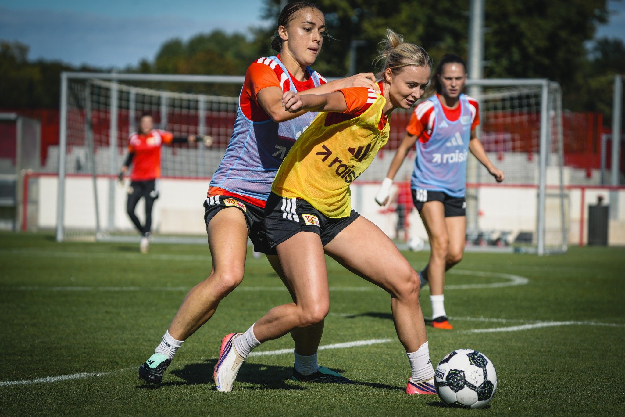 Zwei Frauen spielen Fußball im Training, eine verteidigt, während die andere den Ball führt. Hintergrund mit Toren und weiteren Spielerinnen.