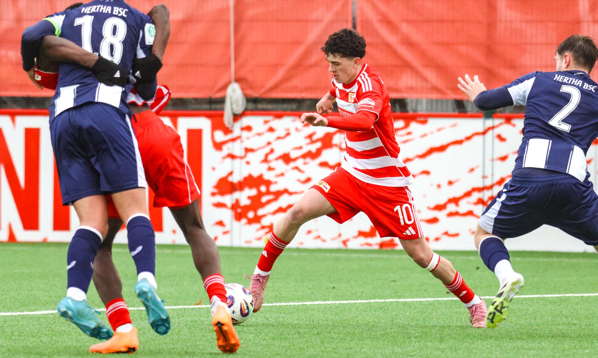 Spielszene mit einem Spieler in roter und weißer Uniform, der zwischen zwei Gegenspielern hindurch dribbelt.