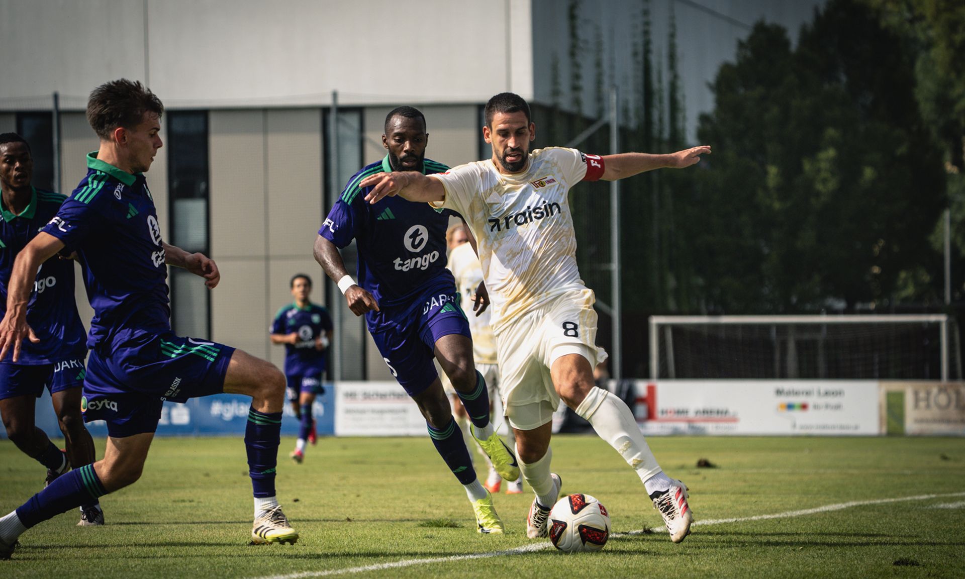 Ein Fußballspieler im weißen Trikot dribbelt den Ball, während zwei Gegner ihn verfolgen. Hintergründe mit Fussballplätzen und Bäumen.