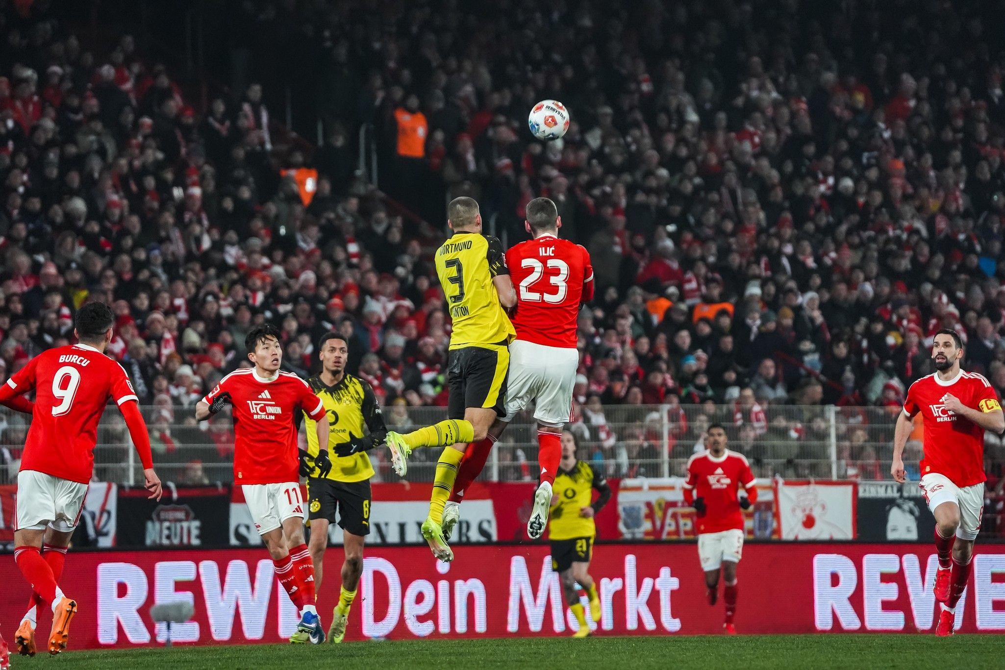 Spielszene aus einem Fußballspiel: Ein Torwart springt hoch, während Spieler anderer Mannschaften im Vordergrund stehen. Fans im Hintergrund.