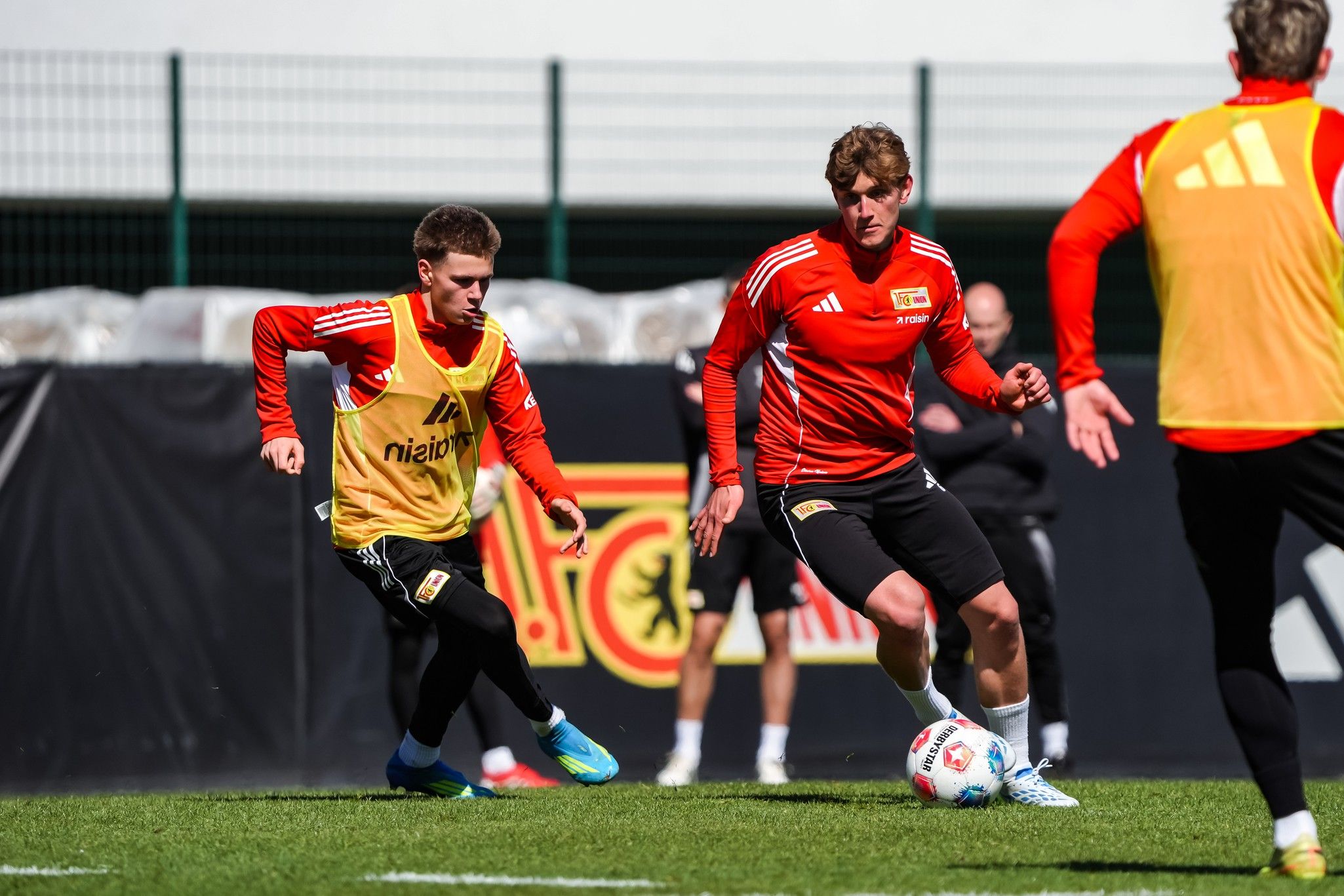 Zwei Fußballspieler im Training, einer mit gelbem Weste, der andere in rotem Trikot, auf einem Fußballfeld.