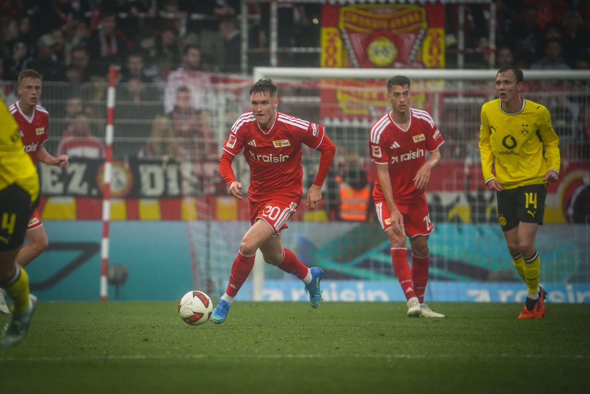 Ein Spieler in rotem Trikot dribbelt den Ball, während andere Spieler und Fans im Hintergrund sichtbar sind.