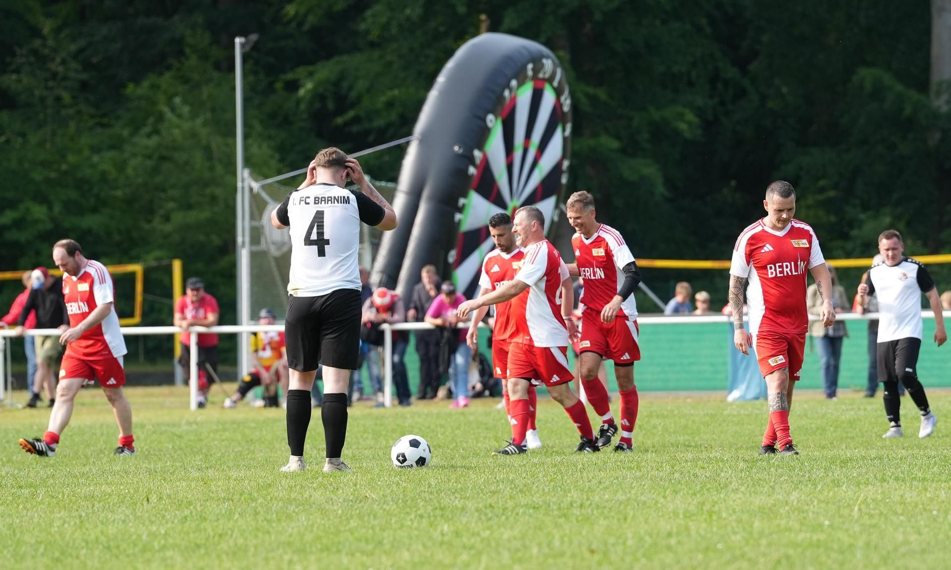 Fußballspiel auf einem Rasenplatz mit Spielern in rot und schwarz, Fans im Hintergrund und einer Dart-Anlage.