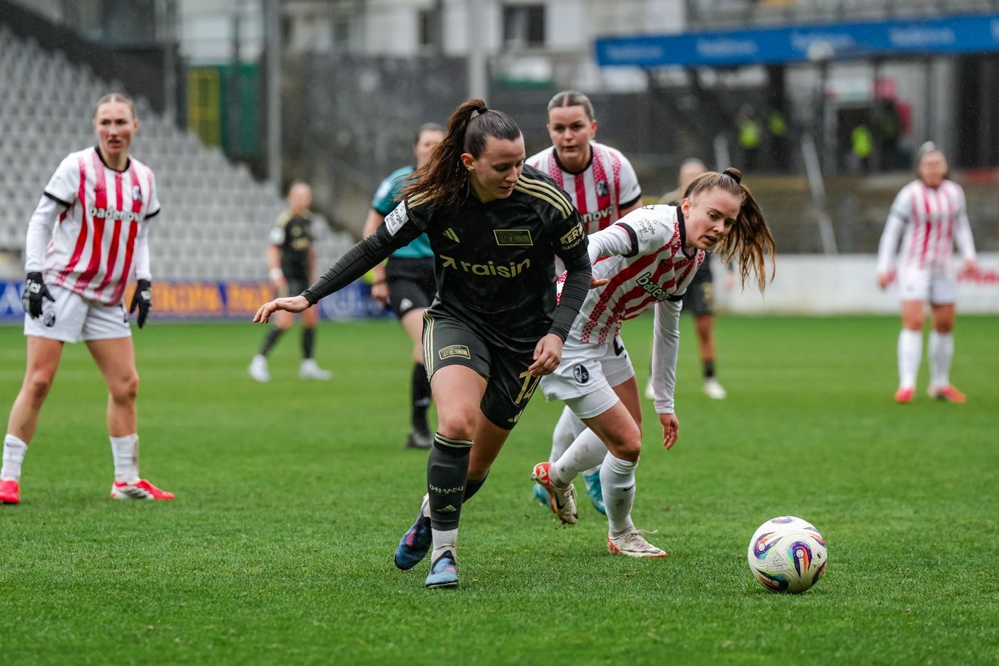 Zwei Fußballspielerinnen kämpfen um den Ball auf einem grünen Spielfeld, im Hintergrund weitere Spielerinnen.