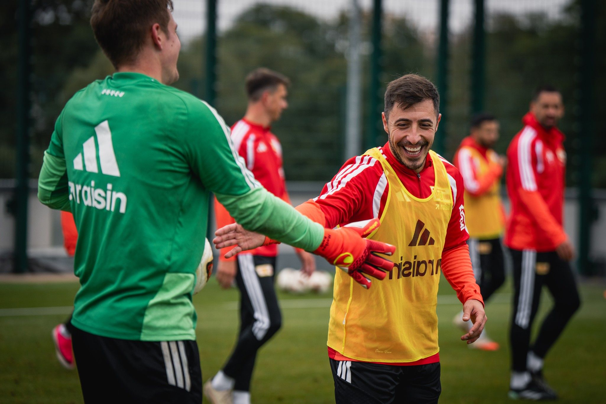 Zwei Fußballspieler lachen und begrüßen sich während eines Trainings auf dem Platz, im Hintergrund weitere Spieler.