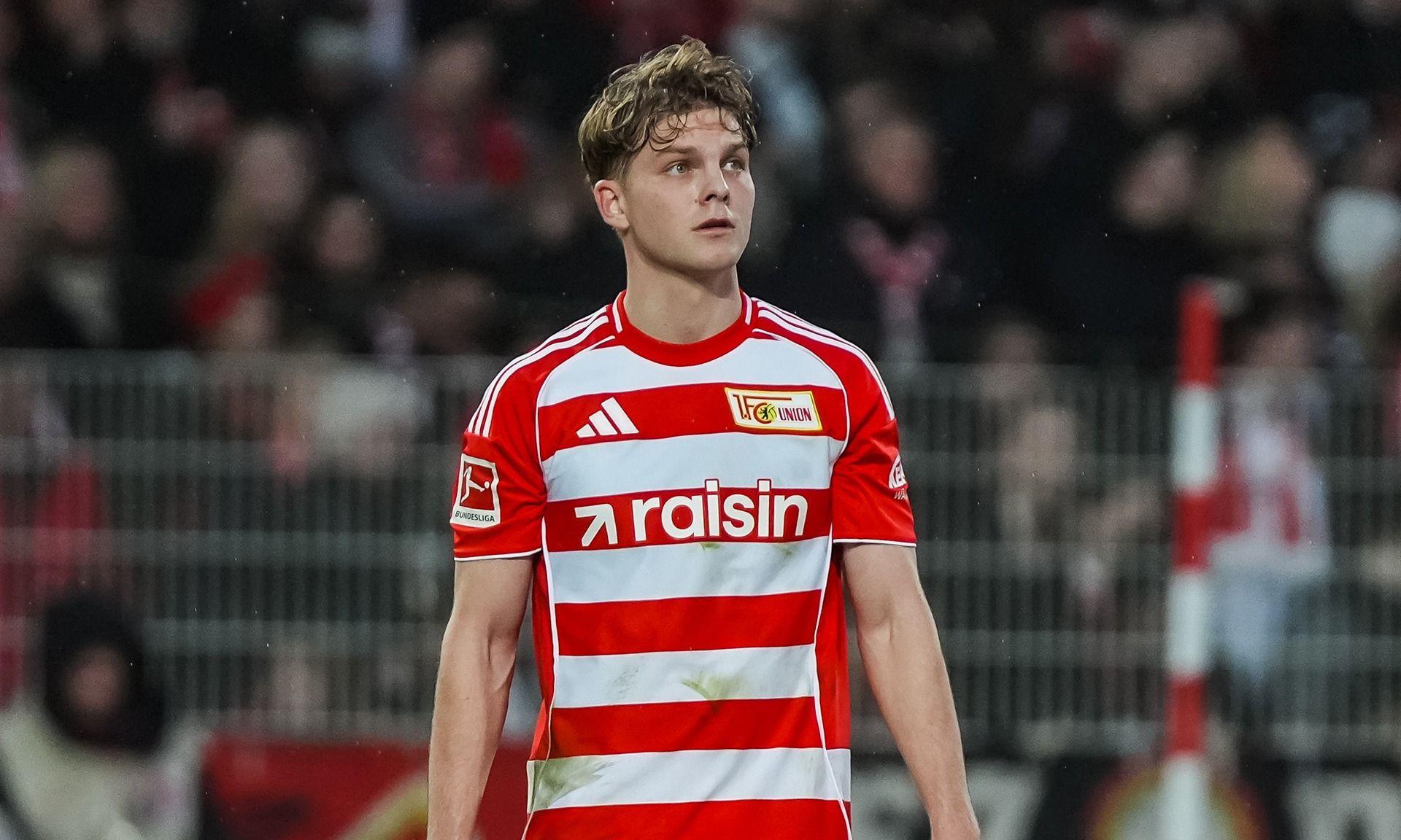 Ein Fußballspieler in einem rot-weiß gestreiften Trikot steht auf dem Spielfeld und schaut nachdenklich.