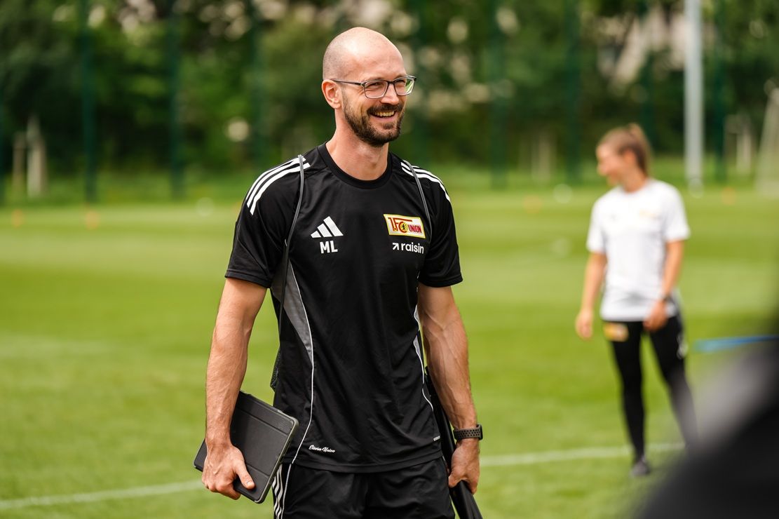 Ein lächelnder Trainer in einem schwarzen Sportoutfit hält ein Tablet in der Hand, während er auf einem Fußballfeld steht.
