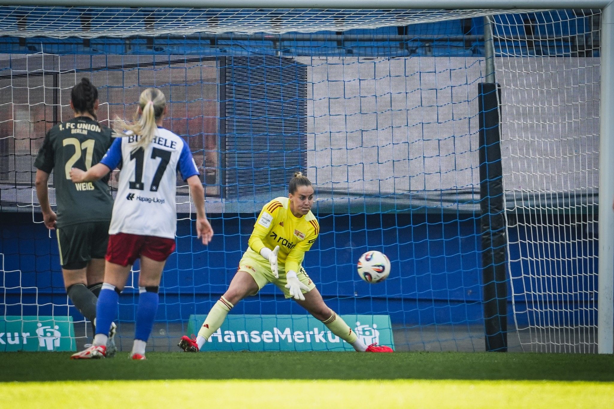 Torhüterin in gelber Ausrüstung weicht einem Schuss aus, während Spielerinnen im Hintergrund im Fußballtrikot agieren.