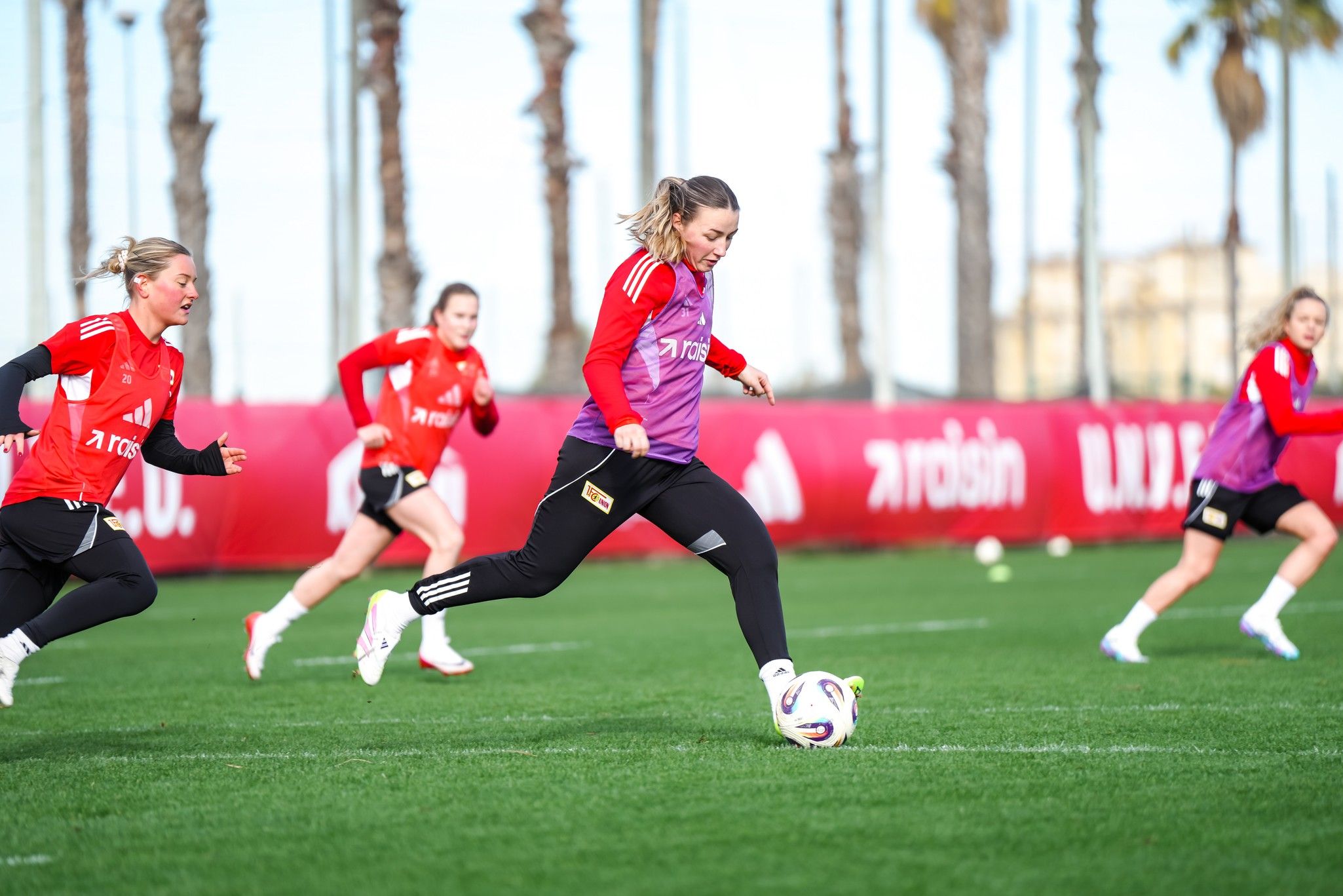 Eine Fußballspielerin tritt den Ball während des Trainings, umgeben von Teamkolleginnen auf einem Rasenplatz. Palmen im Hintergrund.