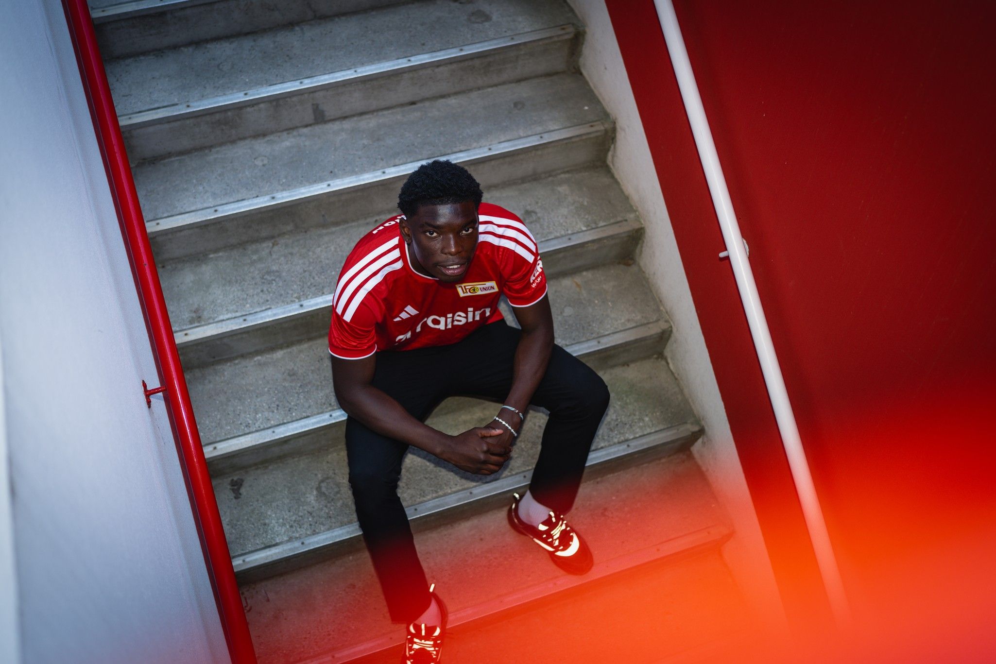 Ein junger Mann in einem roten Sporttrikot sitzt auf einer Treppe und schaut in die Kamera. Die Umgebung ist grau und rot.