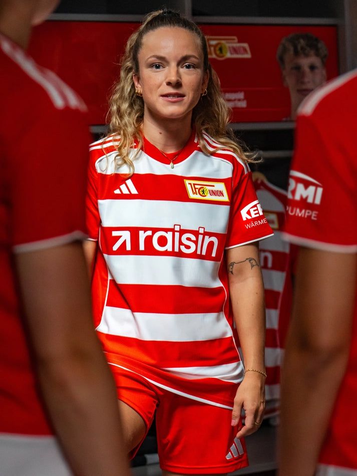 Eine Spielerin in einem rot-weiß gestreiften Fußballtrikot steht in einer Umkleidekabine.
