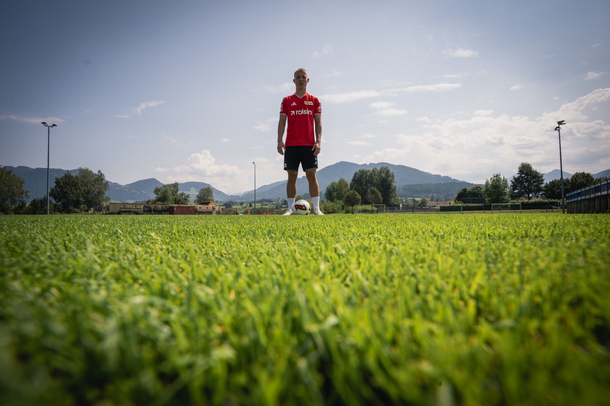 Ein Fußballspieler steht auf einem Rasensportplatz vor Bergen und strahlend blauem Himmel.