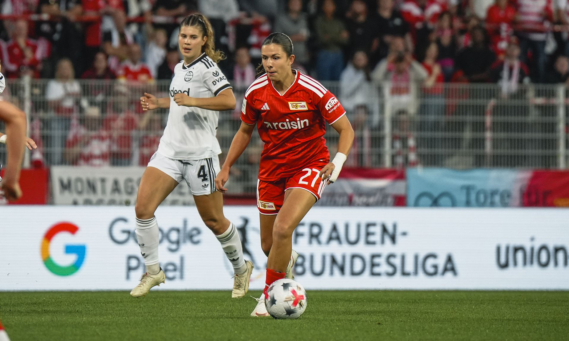Spielszene aus einem Fußballspiel, in dem eine Spielerin in rotem Trikot den Ball führt, während eine Gegenspielerin in Weiss verfolgt.