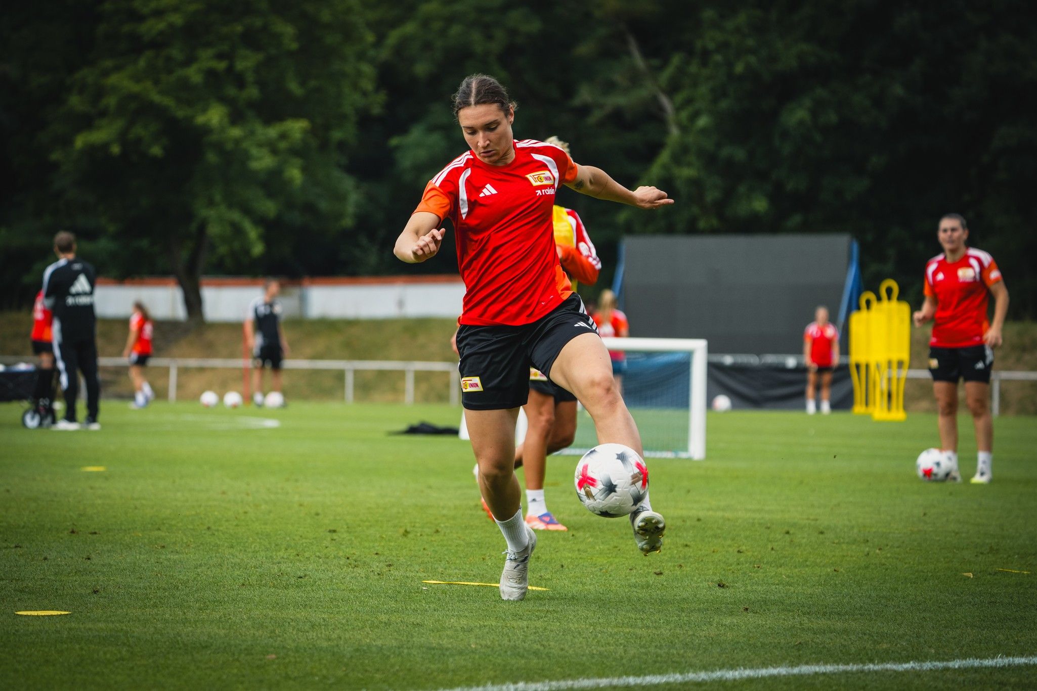 Spielerin in rotem Trikot, die im Training einen Fußball mit dem rechten Fuß kickt, auf einem grünen Platz.