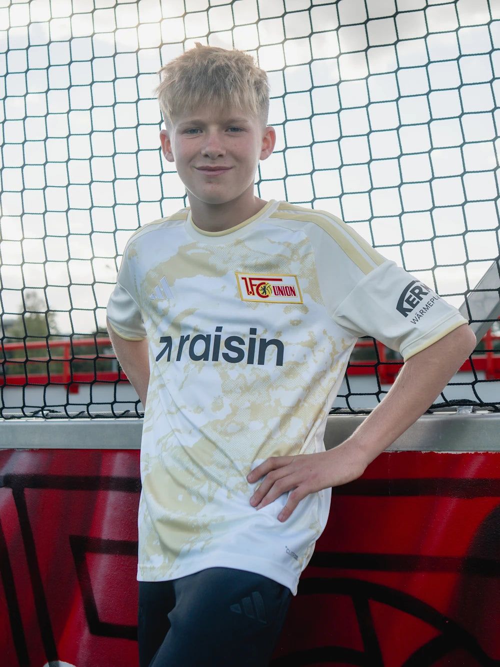Junger Fußballspieler in einem weißen Trikot mit Goldakzenten steht vor einem Fußballnetz und posiert lächelnd.