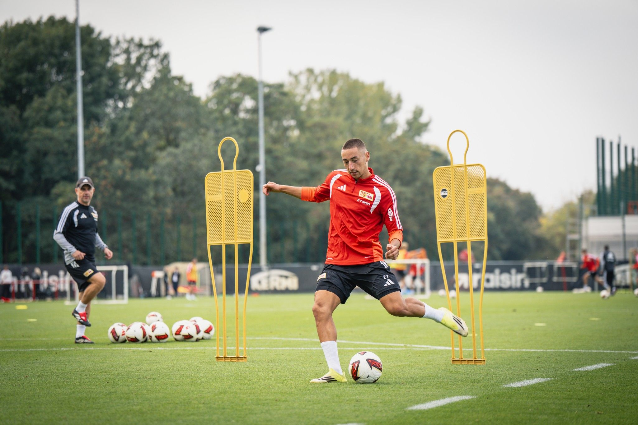 Ein Fußballspieler in roter Trainingsbekleidung schießt auf ein Tor. Im Hintergrund sind Trainingshilfen und mehrere Fußballbälle zu sehen.